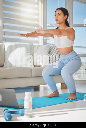 Beauty may be skin deep, but fit is to the bone. a sporty young woman using a laptop and doing squats while exercising at home. Stock Photo