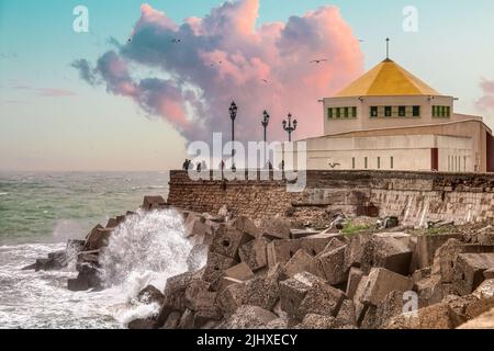 People contemplating the waves breaking against the rocks from a bastion on the La Caleta promenade, with a dramatic sky, in Cádiz, Andalusia, Spain Stock Photo
