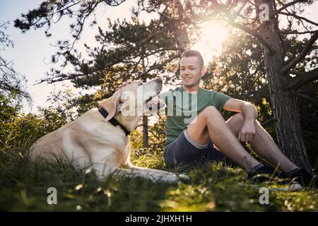 Man with his dog sitting in grass under tree. Pet owner stroking his labrador retriever during sunny summer day. Stock Photo