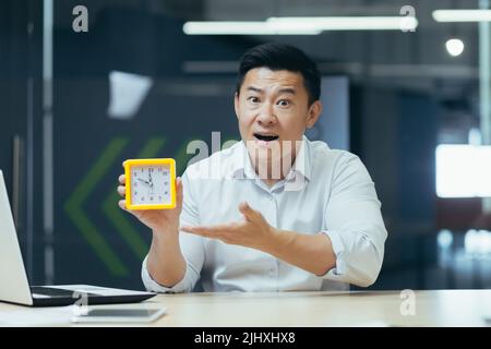 Tired and overworked asian businessman working late in office, holding clock, waiting for decision result Stock Photo
