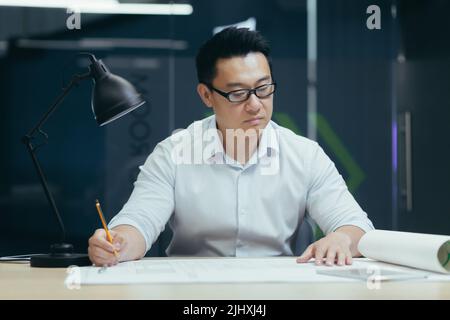 Asian designer working on blueprint in modern office, male architect thoughtfully working on construction project Stock Photo