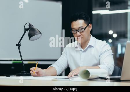 Asian designer working on blueprint in modern office, male architect thoughtfully working on construction project Stock Photo