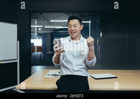 Successful financier businessman asian standing in office, reading good news from phone, holding hand up, celebrating victory and triumph Stock Photo