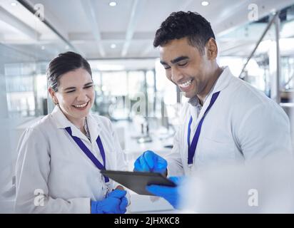 Two happy and cheerful scientists laughing and using a digital tablet while working together in a lab. Mixed race man and caucasian woman checking Stock Photo
