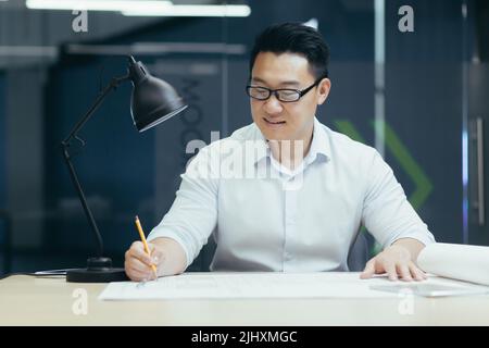 Successful Asian architect works in design studio, on plan drawing of project, makes corrections Stock Photo