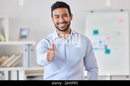 Young happy handsome mixed race businessman showing a thumbs up standing in an office alone at work. One pleased hispanic male boss smiling expressing Stock Photo