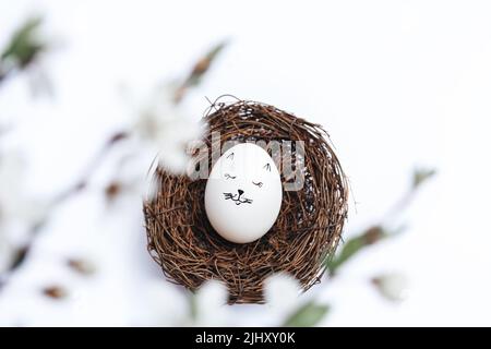 Happy Easter. Beautiful white egg in wicker nest on white background. Picture of gentle kitten. Top view through blurred blooming spring branches. Stylish gentle spring template with Copy Space Stock Photo