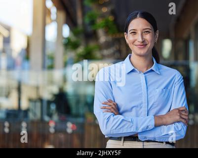 Portrait of a cheerful mixed race smiling businesswoman making with arms folded outside. Young hispanic woman business owner standing outside against Stock Photo