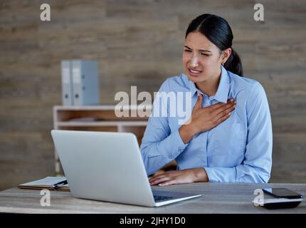 Young hispanic woman suffering from chest pain in office. Mixed race businesswoman feeling unwell while using a laptop at work Stock Photo