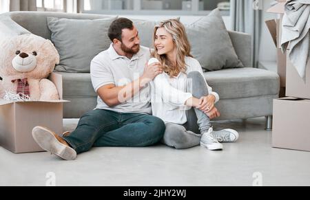 Loving couple looking at each other while sitting in their new home. Young married couple looking happy while sitting on living room floor in their Stock Photo