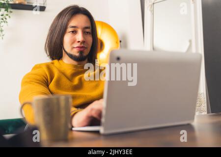 A young adult man with black long hair and a beard wearing an orange sweater working at home office. High quality photo Stock Photo