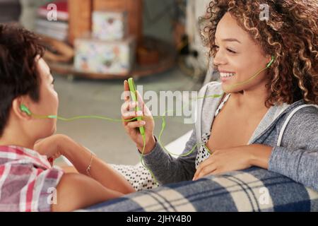 We have the same taste in music. two friends listening to music on a cellphone while sitting on the couch at home. Stock Photo