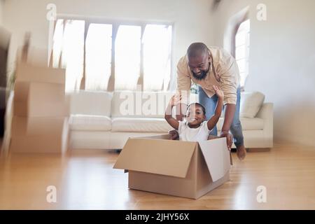 Cheerful african american father pushing excited little girl around in carton box, riding around and having fun in living room. Happy dad and daughter Stock Photo