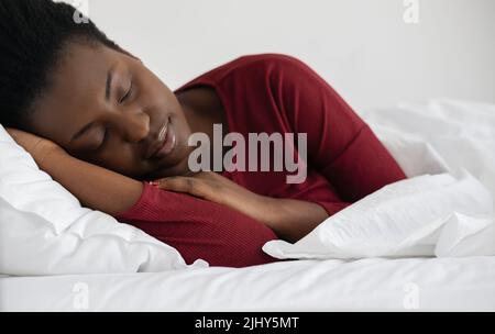 Young beautiful African woman sleeping in white linen bed, wearing red pajamas. Stock Photo
