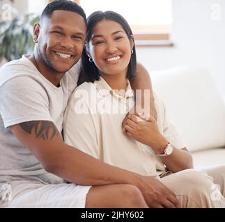 Portrait of smiling mixed race couple sitting together at home. Loving and affectionate man sitting with his arm around his wife relaxing on the couch Stock Photo