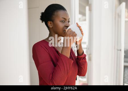 Portrait of a beautiful African woman standing in door way of home, holding a mug, looking into distance Stock Photo