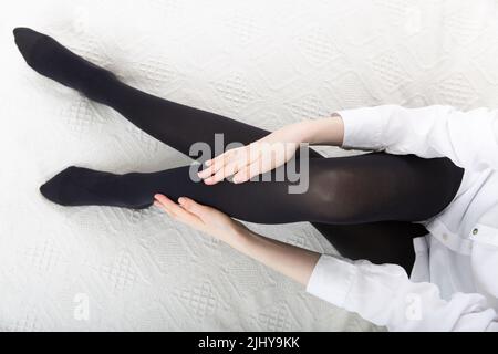 Girl putting on stockings at home. Black compression stockings on a woman in a white room. Black tights. Beautiful female legs Stock Photo