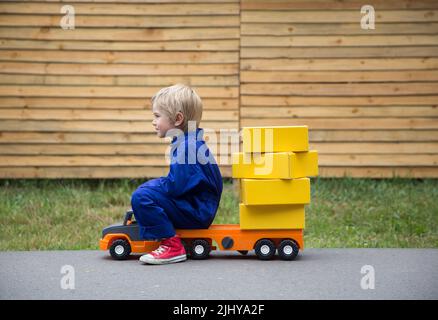 6-year-old boy in a blue overalls - uniform sits on a large toy car - a truck with a lot of yellow cardboard boxes. Parcel delivery, postman, little t Stock Photo