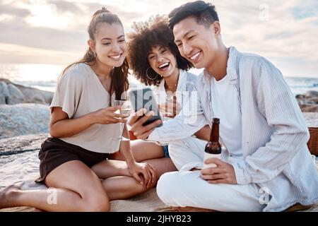 Three friends relaxing and using a phone while sitting and having alcohol at the beach Stock Photo