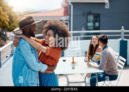 Happy interracial couple hugging while spending time together outside at a restaurant with friends. African american man and mixed race woman Stock Photo
