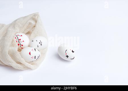Easter beautifully painted eggs in ecological mesh bag on white table. Funny faces, flowers, gentle beauty with red lips, cat's face are painted on eggs. Handmade master class. Soft focus. Copy space Stock Photo