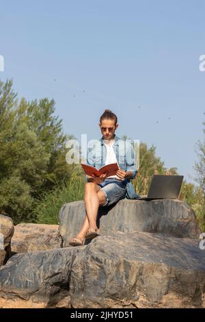 Low angle portrait of a university student reading a magazine next to his laptop into de woods with copy space Stock Photo