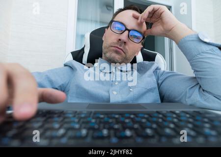Handsome serious man sitting at the office desk with keyboard and eyeglasses against white wall in modern office Stock Photo