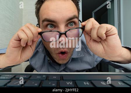 The young surprised man with his laptop computer. An employee in the office looks at the screen in surprise. Crazy programmer at work. Stupid nerd is Stock Photo