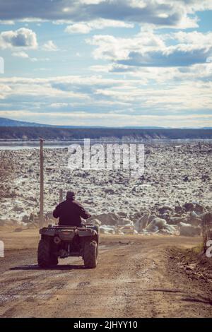 Man driving an ATV on dirt road in northern Indigenous community of Tulita, with Great Bear River breaking up in spring, Northwest Territories, Canada Stock Photo