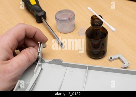 view of the hand of a service center master who glues a torn piece of plastic to a part from a printer on a wooden table Stock Photo