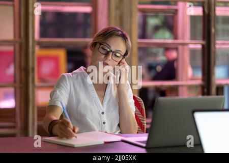 Busy young woman talking on smartphone, making notes while studying in public library with laptop Stock Photo