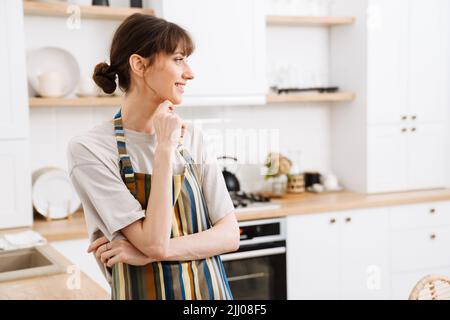 White young woman wearing apron smiling while standing in kitchen at home Stock Photo