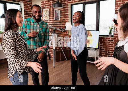 Laughing friends in living room at home having fun together while listening to funky disco music. Joyful group of multiethnic people dancing and celebrating birthday at wine party event. Stock Photo