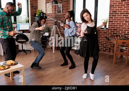 Diverse group of close friends dancing in living room while celebrating birthday event. Multicultural people relaxing with music while enjoying wine party at home together. Stock Photo