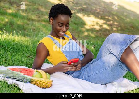 Very happy smiling black African woman lying in an outdoor park chatting and texting on her cell phone while having a picnic  Stock Photo