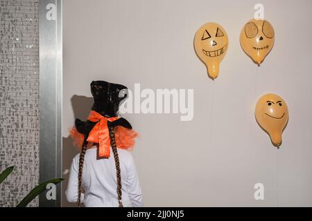 Back view Child in fancy dress for happy halloween, home party celebration. Hat with red wig on head of girl with long braided braids. Orange balls on wall. Celebration of Day of All Saints and Dead. Stock Photo