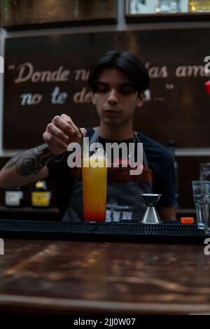 Bartender garnished a freshly brewed yellow drink with a green leaf. Waiter working at the bar with a drink for a client. Latino barman preparing a glass of juice Stock Photo