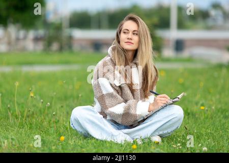 A young woman writer or student with a notebook is sitting on the lawn. She looks away thoughtfully. Stock Photo