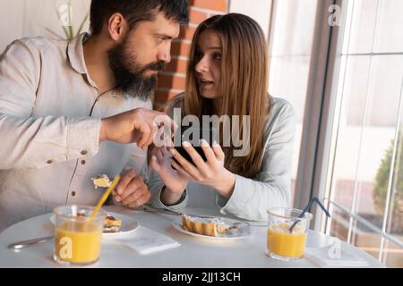 Portrait of happy cute young couple looking at phone and smiling while eating and sitting in a cafe. Joyful couple using cellphones and laughing while Stock Photo