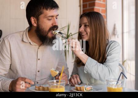 Young couple eating meal at home, loving woman feeding her boyfriend. Lovers having tasty helthy breakfast in kitchen, enjoying spending morning time Stock Photo