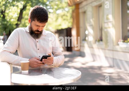 Serious male journalist having rest while drinking coffee while texting with friends on mobile phone at city street Stock Photo