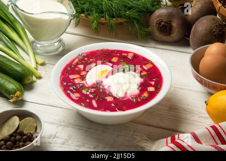 Traditional Ukrainian summer cold borscht with beetroot, cucumbers, boiled egg in a ceramic bowl with ingredients on a white old wooden rural table. Stock Photo