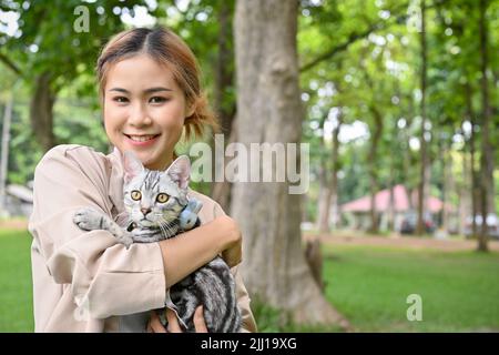 Beautiful and attractive young Asian woman holds a little cute cat in the green city park. Outdoor activity with an animal concept Stock Photo