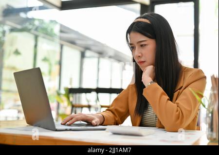 Thoughtful young Asian businesswoman or office employee concentrated focused working on her assignment project, using laptop computer. Stock Photo