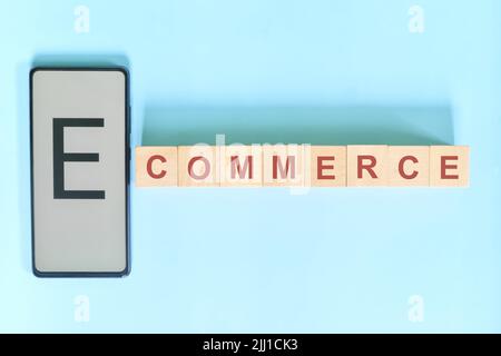 E-commerce business concept. Hand holding mobile phone with word on wooden blocks flat lay in blue background. Stock Photo