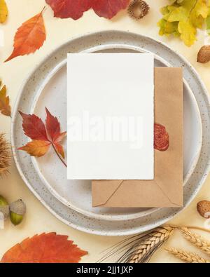 Autumn table place with colorful leaves and blank card with envelope  top view. Mockup of fall vertical card on light yellowtable, flat lay, copy spac Stock Photo