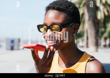 cheerful african woman sending voice message outdoors on a sunny day. She wears casual summer attire and smiles happily Stock Photo