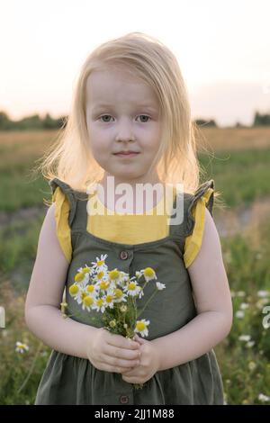 A little blonde girl is sitting walking on a chamomile field and collecting a bouquet of flowers. The concept of walking in nature, freedom and a clea Stock Photo