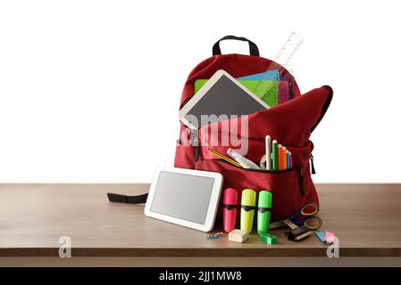 School backpack full of school supplies on wooden table with electronic books and white isolated background. Front view. Stock Photo