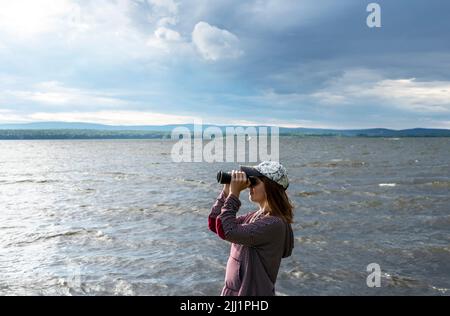 Young woman in cap looking through binoculars at birds on the lake or river Birdwatching, zoology, ecology Research in nature, observation of animals Stock Photo